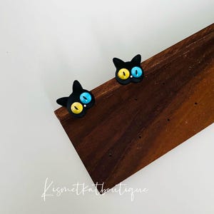 May include: A pair of black cat earrings with blue and yellow eyes. The earrings are on a wooden surface.