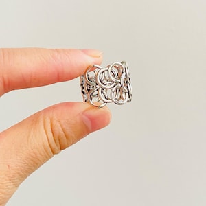 May include: A silver ring featuring a wide band constructed from multiple interlocking circular links. The metal has a polished finish with visible dark oxidation in the recessed areas between the connected rings, creating a textured appearance.