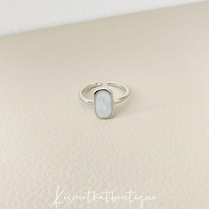 Könnte beinhalten: Ein silberner Ring mit einem weißen ovalen Stein. Der Ring liegt auf einem weißen Hintergrund.