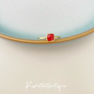 May include: A gold-colored ring with a square, bright red gemstone. The ring is set on a light blue plate with a brown rim, against a white background. The ring's design is simple and elegant, showcasing the vibrant red stone.