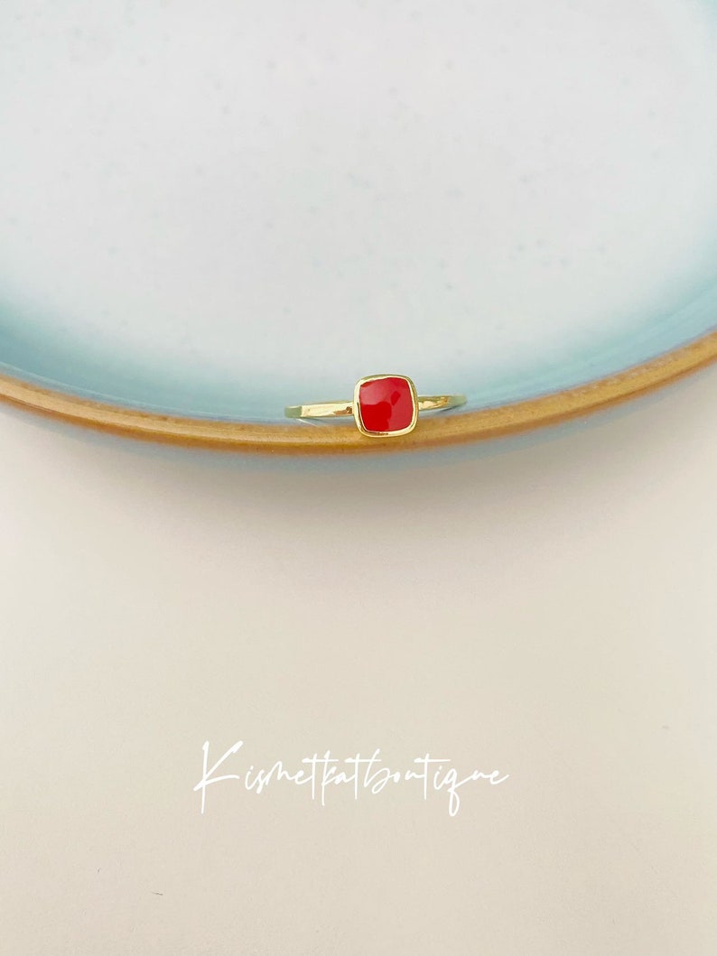 May include: A gold-colored ring with a square, bright red gemstone. The ring is set on a light blue plate with a brown rim, against a white background. The ring's design is simple and elegant, showcasing the vibrant red stone.
