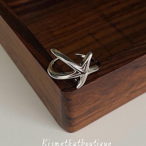 May include: A polished silver-toned ring featuring a five-pointed starfish design with a smooth, reflective surface. The band is open-ended, allowing for adjustable sizing. The text Kismetkatboutique is printed in white below the ring.