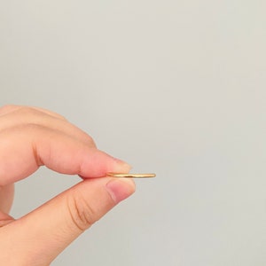 May include: A thin gold band ring held between two fingers.