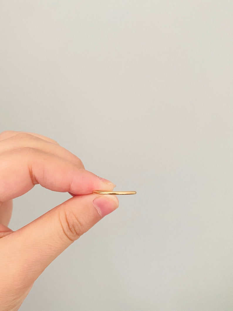 May include: A thin gold band ring held between two fingers.