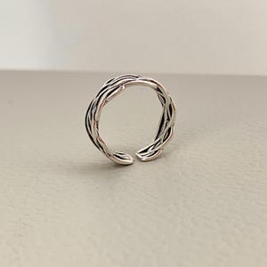 以下が含まれることがあります： 編み込みデザインのシルバーカラーのリング。リングは前面が開いており、テクスチャパターンがあります。「Kismetkatboutique」の文字が画像の下部にあります。
