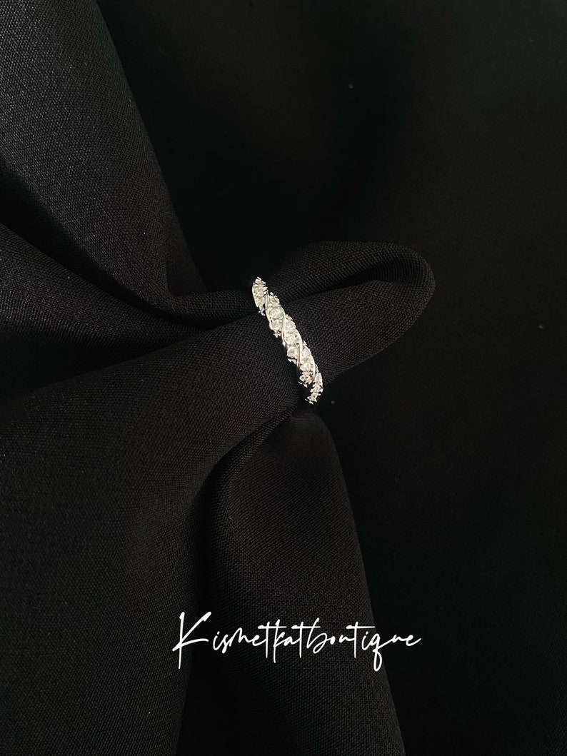 Op de afbeelding: Een zilveren ring met een doorlopende rij kleine, sprankelende stenen. De ring is geplaatst tegen een zwarte stoffen achtergrond. De tekst "Kismetkatboutique" staat in cursief onderaan de afbeelding.
