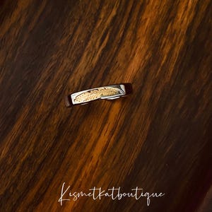 May include: A silver ring with a gold feather design. The ring is on a dark wood surface.