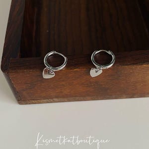 May include: A pair of silver hoop earrings with a small heart charm hanging from each hoop.