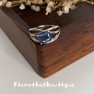 Puede incluir: Un anillo de tono plateado con una piedra preciosa azul rectangular engastada en un diseño de banda entrecruzada. El anillo se muestra con el texto Kismetkatboutique visible debajo. La banda tiene una estructura abierta y ajustable.