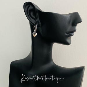 May include: Silver hoop earrings with a small heart charm dangling from each hoop.