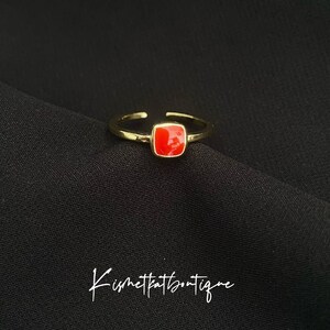 May include: A gold-toned adjustable ring with a square, bright red stone. The ring is set against a black background. The text "Kismetkatboutique" is written in cursive at the bottom of the image.