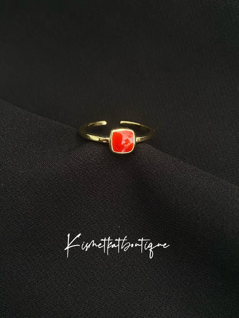 May include: A gold-toned adjustable ring with a square, bright red stone. The ring is set against a black background. The text "Kismetkatboutique" is written in cursive at the bottom of the image.
