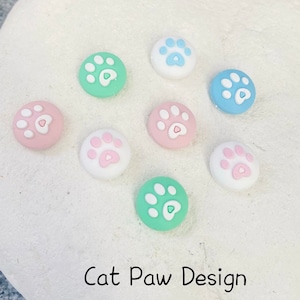 以下が含まれることがあります： ピンクのハートが中央に描かれた、カラフルな猫の足の形をしたボタンが8つあります。ボタンは白い表面に円形に配置されています。ボタンの下には「Cat Paw Design」という文字が書かれています。
