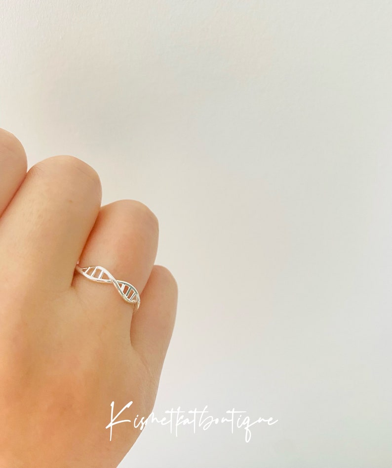 May include: A silver ring featuring a DNA double helix design. The ring is displayed on a finger against a white backdrop. The ring's design is a delicate representation of the genetic structure. The image includes the text "Kismetkatboutique".