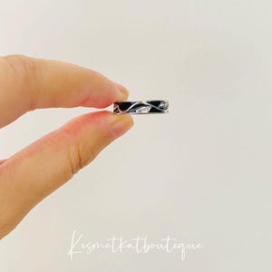 May include: A silver ring with a black enamel band and a delicate vine design. The vine is accented with small white stones.