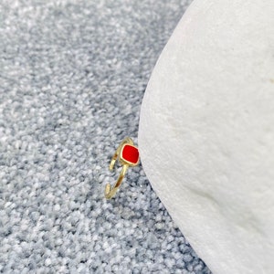 May include: A gold-colored ring with a square, red enamel center stone. The ring is set against a gray, textured background and a white object. The ring's band is open and adjustable.