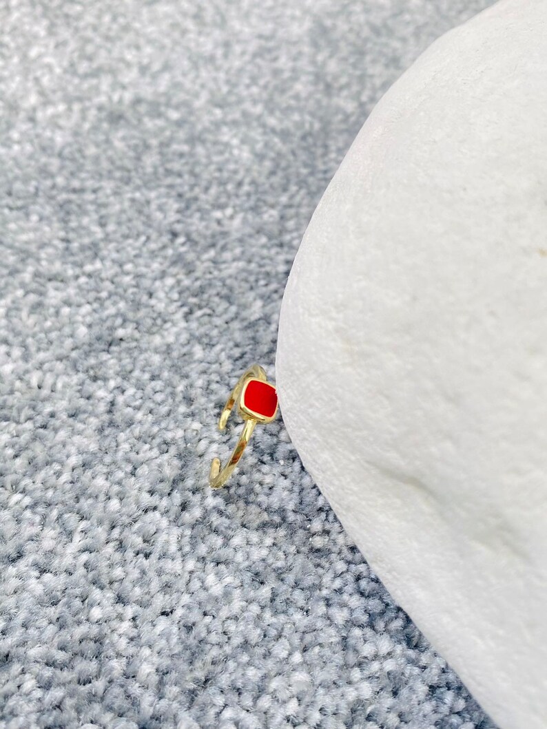 May include: A gold-colored ring with a square, red enamel center stone. The ring is set against a gray, textured background and a white object. The ring's band is open and adjustable.