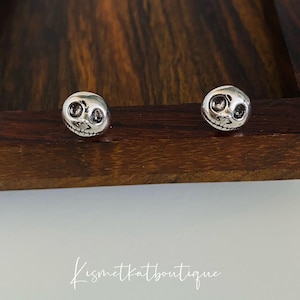 May include: A pair of silver earrings featuring a detailed skull design. The earrings are displayed on a wooden surface.