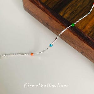 May include: A delicate silver bracelet with colorful beads. The bracelet features small, round beads in red, black, green, blue, and orange, interspersed along a silver chain. The bracelet is displayed on a wooden surface with the text "Kismetkatboutique".