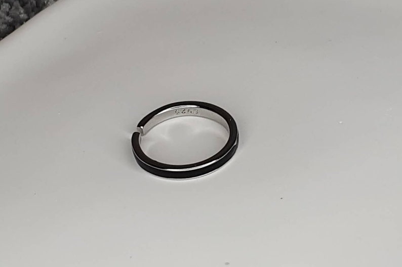 Pu&ograve; includere: Un anello d'argento con una fascia nera attorno al centro. L'anello ha il numero "6243" inciso all'interno.