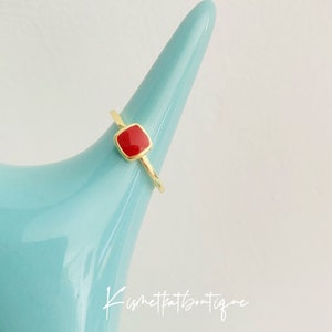 May include: A gold-colored ring with a square, red gemstone. The ring is displayed on a light blue, cone-shaped object. The ring's band is thin and delicate. The gemstone is set in a gold bezel. The ring is a simple, elegant piece of jewelry.