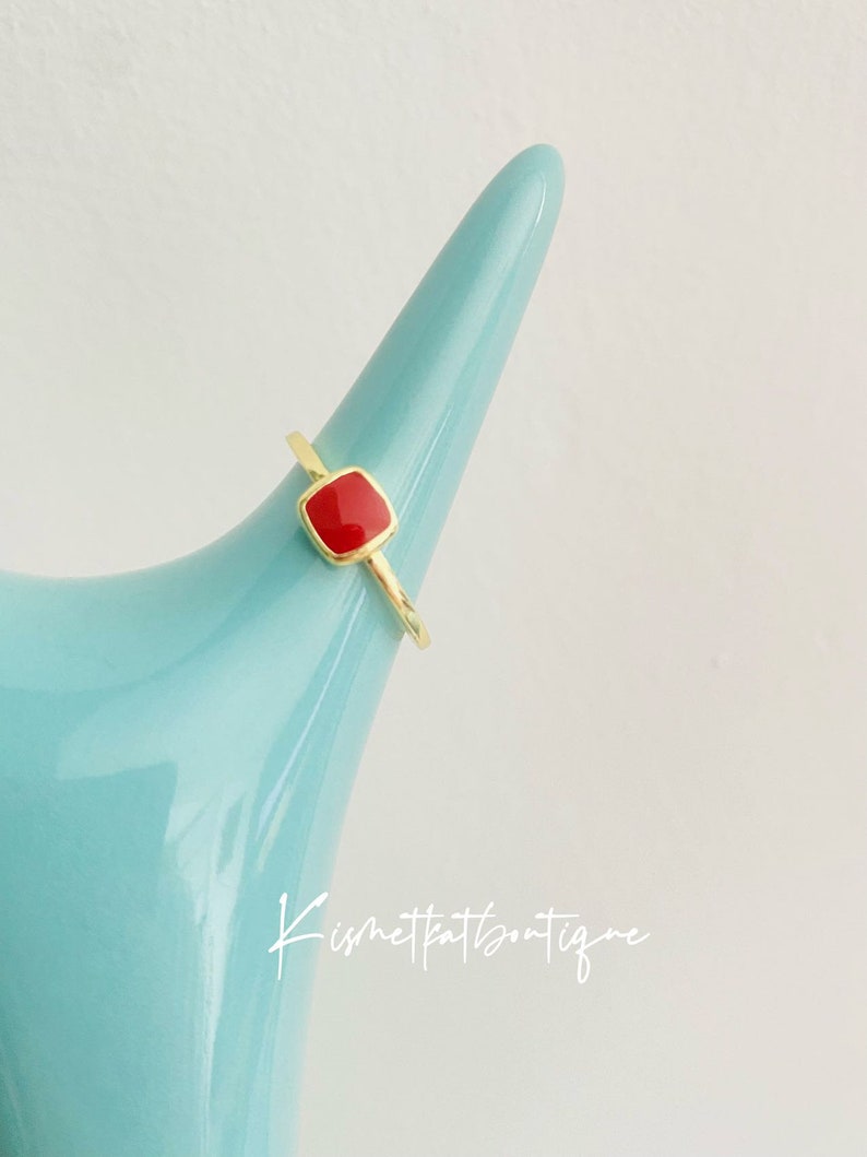 May include: A gold-colored ring with a square, red gemstone. The ring is displayed on a light blue, cone-shaped object. The ring's band is thin and delicate. The gemstone is set in a gold bezel. The ring is a simple, elegant piece of jewelry.