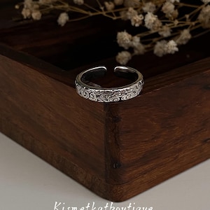 Könnte beinhalten: Verstellbarer Ring in Silber mit eingraviertem Wirbelmuster. Der Ring wird auf einer dunklen Holzoberfläche mit getrockneten Blumen im Hintergrund präsentiert. Der Shop-Name "Kismetkatboutique" ist sichtbar.