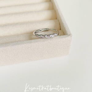 Op de afbeelding: Een zilverkleurige ring met een gevlochten ontwerp, bezet met kleine, heldere edelstenen. De ring wordt gepresenteerd op een lichtgekleurd, getextureerd oppervlak. De merknaam "Kismetkatboutique" staat onderaan in cursief.