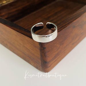 May include: A silver-colored adjustable ring with a flat band. The ring is open at the top and sits on a wooden box. The ring is a simple, modern design.
