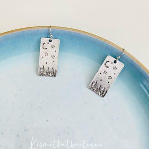 Op de afbeelding: Een paar zilveren oorbellen met een rechthoekig ontwerp met een halve maan, sterren en een silhouet van een bos.