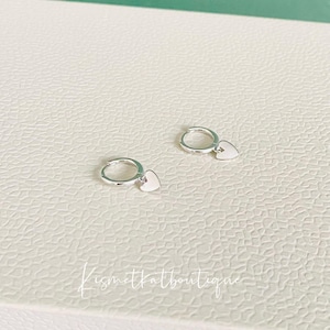 May include: A pair of silver hoop earrings with a small heart charm hanging from each hoop.