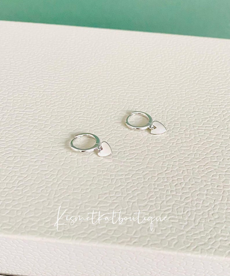May include: A pair of silver hoop earrings with a small heart charm hanging from each hoop.