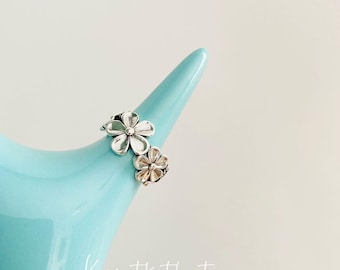 May include: Two silver flower rings displayed on a light blue ceramic object. The rings have detailed petals and are positioned at different heights. The background is a soft, neutral color, highlighting the jewelry.