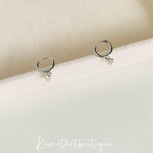 May include: A pair of silver hoop earrings with a small heart charm hanging from each hoop.