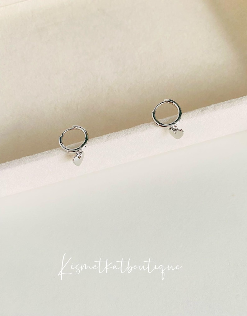 May include: A pair of silver hoop earrings with a small heart charm hanging from each hoop.