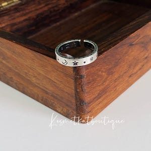 Puede incluir: Un anillo de plata con un diseño de luna y estrellas. El anillo está en una caja de madera.