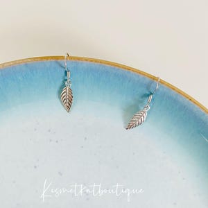 Puede incluir: Un par de pendientes de hoja de plata sobre un plato azul claro. Los pendientes son simples y delicados, con un pequeño dije de hoja colgando de un gancho de plata.