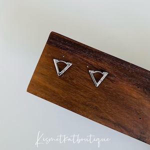 Puede incluir: Un par de pendientes de aro triangulares de plata en un expositor de madera. Los pendientes son de diseño simple y moderno.