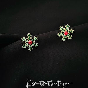 Puede incluir: Un par de pendientes de copo de nieve de oro con esmalte verde y detalles de cristal rojo. Los pendientes están sobre un fondo negro.
