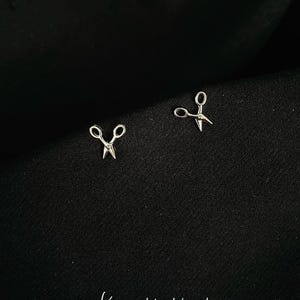 Può includere: Un paio di orecchini a forma di forbici argentati su uno sfondo nero. Gli orecchini sono piccoli e delicati e sono perfetti per aggiungere un tocco di fantasia a qualsiasi outfit.