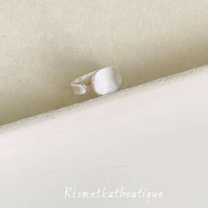 May include: A silver-toned open ring featuring a circular, brushed-finish face and a smooth, adjustable band. The text Kismetkatboutique is visible in the lower portion of the frame, presented in a simple, white, sans-serif font style.