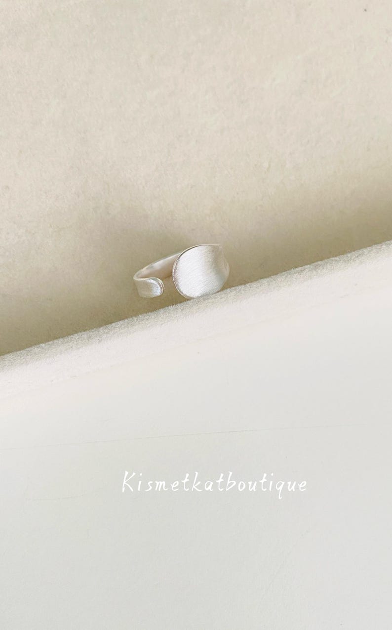 May include: A silver-toned open ring featuring a circular, brushed-finish face and a smooth, adjustable band. The text Kismetkatboutique is visible in the lower portion of the frame, presented in a simple, white, sans-serif font style.
