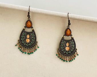 May include: A pair of bronze-toned dangle earrings featuring a teardrop-shaped amber stone at the top, a central arch with green and peach-colored accents, and a fringe of small green and peach beads hanging from the bottom edge.