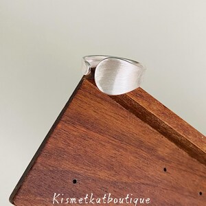 May include: A silver-toned ring with a wide, smooth, brushed metal surface and an open-ended band design. The ring is positioned on a dark brown wooden display stand that features the text Kismetkatboutique printed in white lettering.