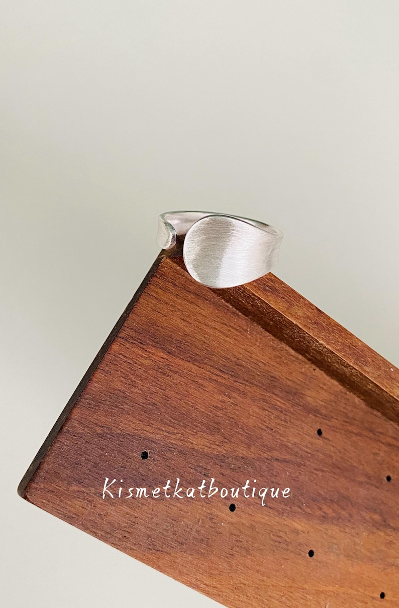 May include: A silver-toned ring with a wide, smooth, brushed metal surface and an open-ended band design. The ring is positioned on a dark brown wooden display stand that features the text Kismetkatboutique printed in white lettering.