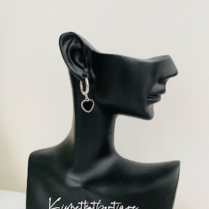 May include: Silver hoop earrings with a black heart charm dangling from the bottom. The earrings are shown on a black mannequin head.