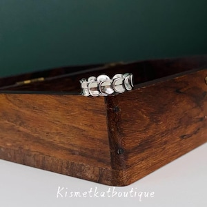 Peut inclure: Une bague en argent avec un motif incurvé répétitif, posée sur une boîte en bois. Le motif de la bague ressemble à une série d'éléments en forme de croissant connectés. La boîte est de couleur marron foncé, et le texte "Kismetkatboutique" est visible.