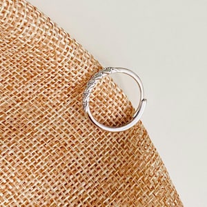 Op de afbeelding: Een zilverkleurige oorring met een decoratief gedeelte van kleine, heldere stenen. De oorring is geplaatst tegen een getextureerde, bruinachtige achtergrond.