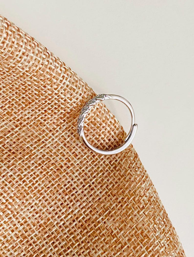 Op de afbeelding: Een zilverkleurige oorring met een decoratief gedeelte van kleine, heldere stenen. De oorring is geplaatst tegen een getextureerde, bruinachtige achtergrond.