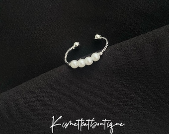 May include: A silver-toned ring with a curved design, featuring five white pearls. The ring is set against a black background. The text "Kismetthatboutique" is written in cursive script.